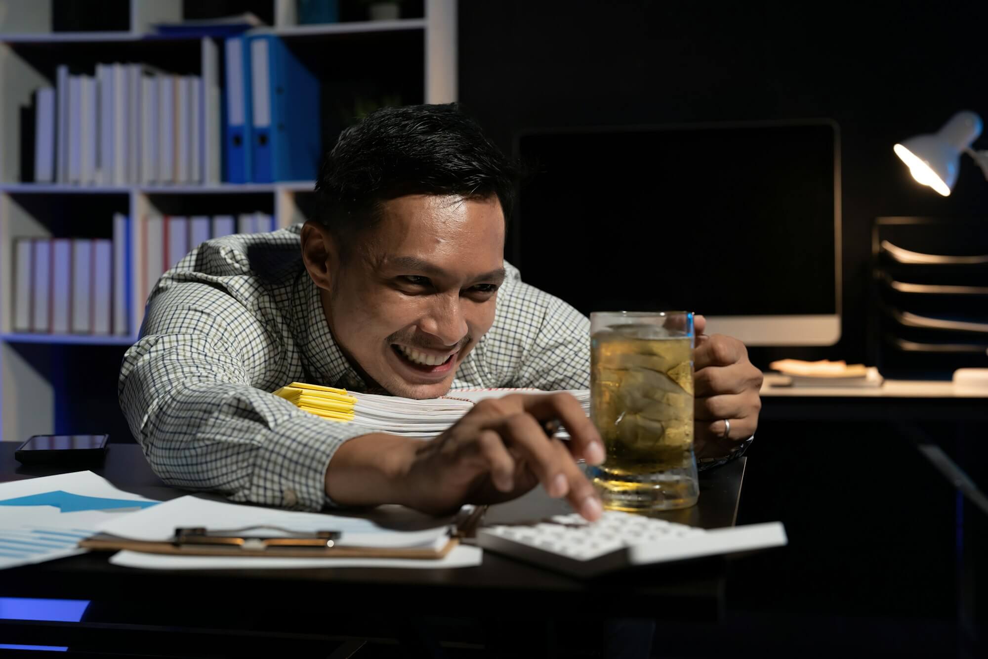 Asian man working overtime drinking beer while using calculator in office at night.