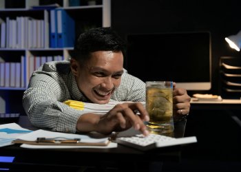 Asian man working overtime drinking beer while using calculator in office at night.